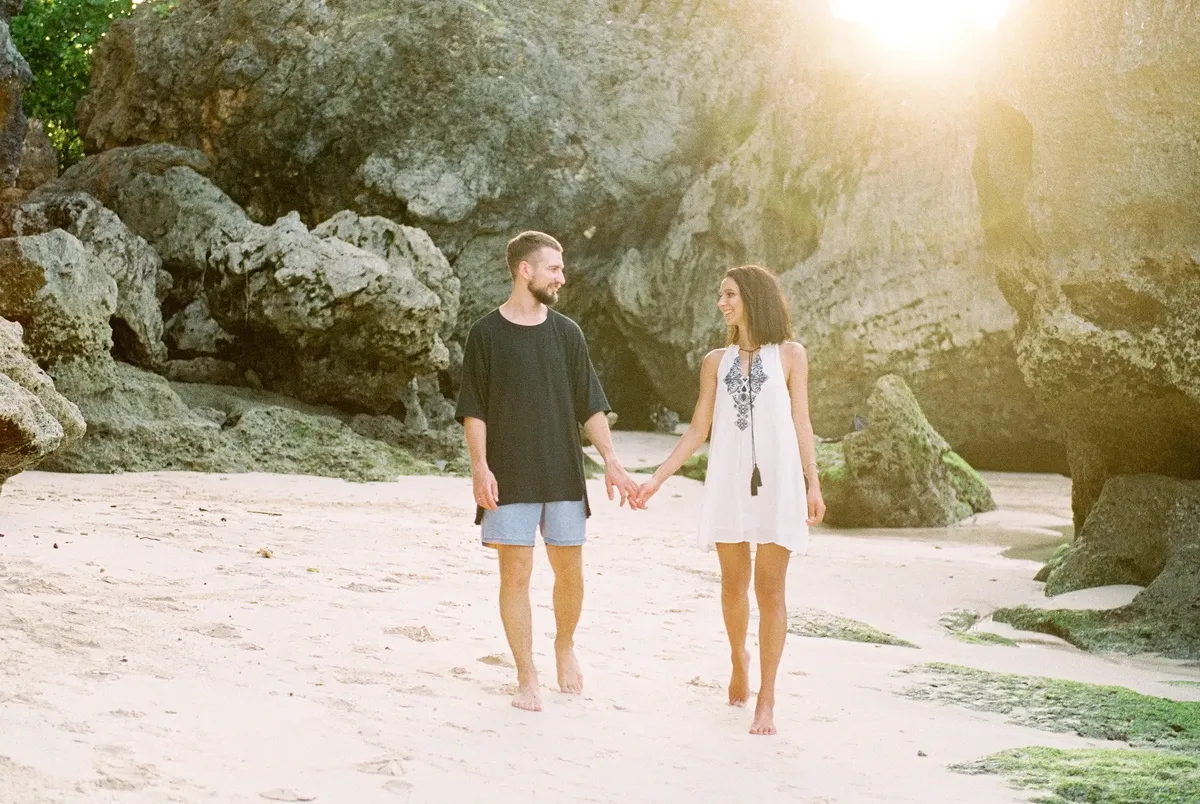 A happy couple holding hands and walking barefoot on a sunny Bali beach.