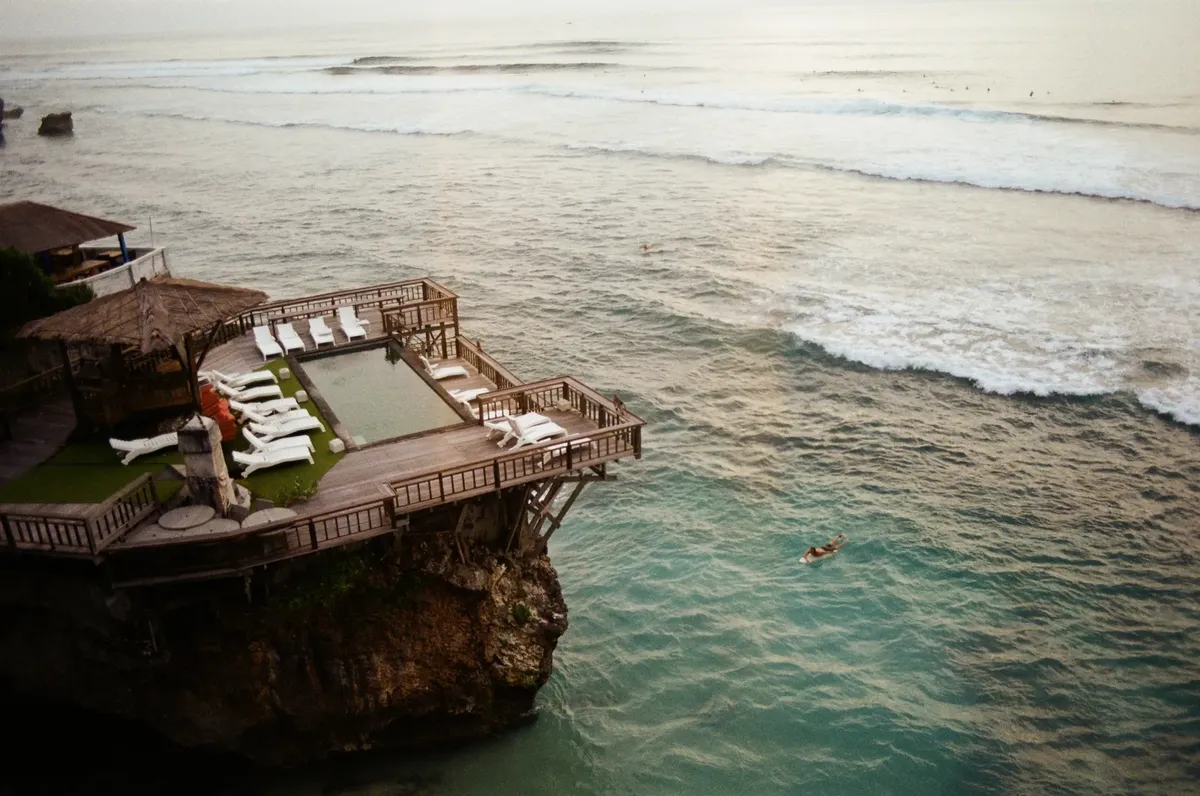 Aerial view of a luxurious cliffside resort in Bali, Indonesia, with ocean and swimmers below.